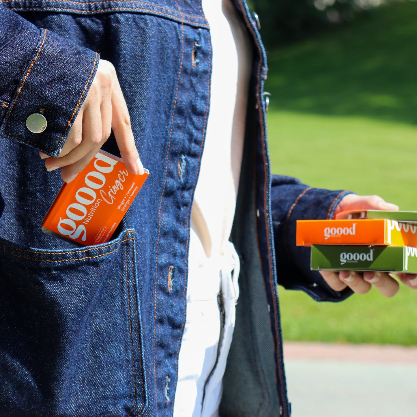 Person carrying Goood Nutrition herbal pastilles outdoors, placing a Goood Ginger box into a denim jacket pocket while holding other Goood Nutrition boxes in hand.