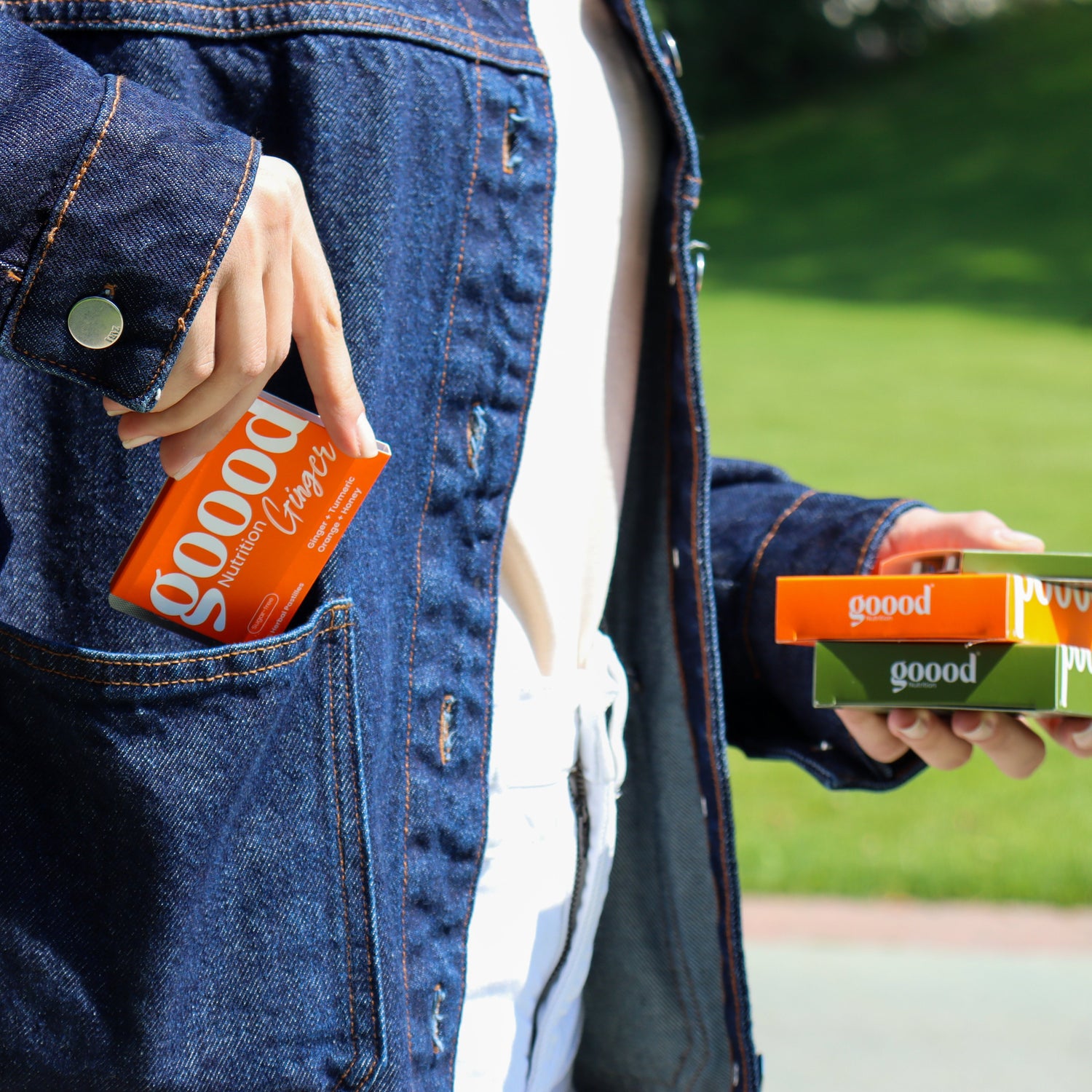 Person carrying Goood Nutrition herbal pastilles outdoors, placing a Goood Ginger box into a denim jacket pocket while holding other Goood Nutrition boxes in hand.